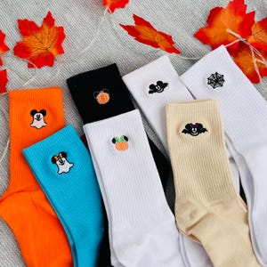 May include: A selection of Halloween-themed socks in a variety of colours. The socks are in shades of orange, blue, black, white, and beige. Each sock has an embroidered Halloween design, including a ghost, a pumpkin, a bat, and a spiderweb. The socks are displayed on a grey surface with autumn leaves.