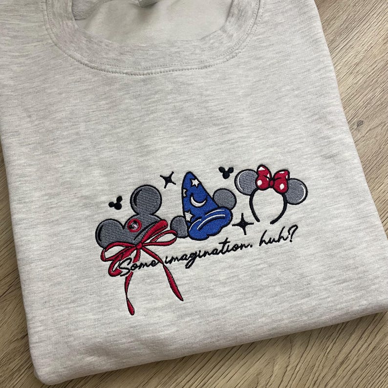 May include: Pale grey sweatshirt with embroidered cartoon character designs. Features a grey mouse head with a red bow, a blue wizard hat, and a Minnie Mouse headband with a red bow. Text reads "Some imagination, huh?"