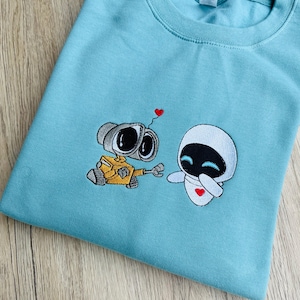 May include: A pale blue sweatshirt with an embroidered design of two cartoon robots, one yellow and grey, the other white. A small red heart floats above them. The sweatshirt is made of a soft material.