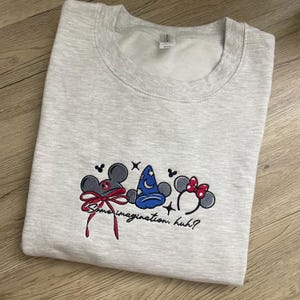 May include: Pale grey sweatshirt featuring embroidered cartoon character ears, a wizard's hat, and a red bow. The phrase "Some imagination, huh?" is embroidered below. The sweatshirt is folded on a wooden surface.
