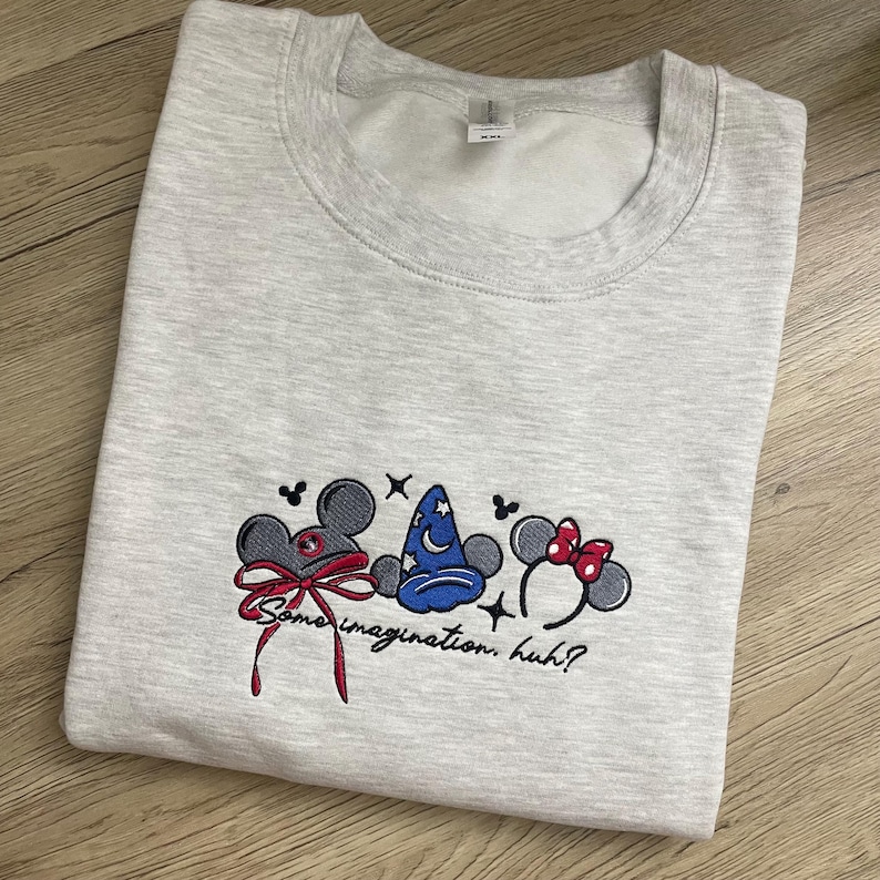 May include: Pale grey sweatshirt featuring embroidered cartoon character ears, a wizard's hat, and a red bow. The phrase "Some imagination, huh?" is embroidered below. The sweatshirt is folded on a wooden surface.