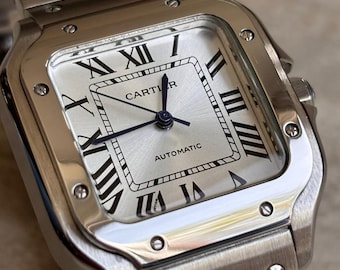 May include: A silver-toned Cartier automatic wristwatch featuring a square case with exposed screws on the bezel and a metallic link bracelet. The silver dial displays black Roman numeral hour markers, blue hands, and the Cartier logo.