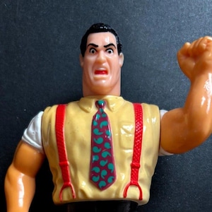May include: A plastic action figure of a man in a yellow shirt, red braces, and a patterned tie. The figure has black trousers and shoes, with one arm raised in a fist.
