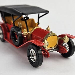 May include: A detailed model of a vintage red car with a black top and black tires. The car features gold accents, a yellow grill, and a tan interior. The model car is a classic collectible.