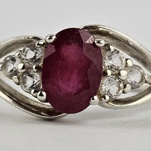 May include: A silver ring featuring a large oval ruby gemstone as the centerpiece. The ruby is a deep red color. Accompanying the ruby are smaller, round, clear gemstones on either side, set in a flowing, elegant design.