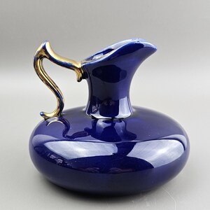 May include: A cobalt blue ceramic pitcher with a gold-colored handle. The pitcher has a rounded body and a spout. The handle is curved and attached to the top of the pitcher. The pitcher is likely for serving beverages.