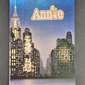 Pode incluir: Uma capa de livreto retangular apresentando uma paisagem urbana noturna de Nova York com arranha-céus iluminados contra um céu azul escuro. A palavra Annie está impressa em letras brancas grandes com contorno marrom, com uma pequena ilustração.