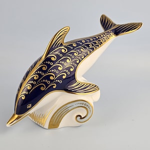 Könnte beinhalten: Eine Delfinfigur aus Keramik mit einem tief dunkelblauen Oberkörper, der mit komplizierten goldenen Wirbelmustern und Punkten verziert ist. Der untere Teil ist weiß mit goldenen Akzenten und zeigt den Delfin, der aus einer Welle springt.