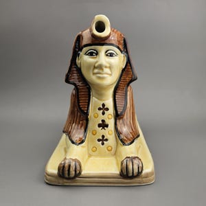 May include: A ceramic Egyptian sphinx figurine in tan, brown, and black. The sphinx has a detailed face, a headdress, and a decorative collar. The figurine is a decorative item, likely for display or collection.