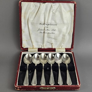 May include: A set of six silver spoons in a red box with a white satin lining. The spoons have a classic design with a rounded bowl and a geometric handle. The box has the text "With Compliments from James Walker Ltd., The Lelen Jewellers."