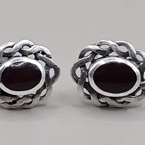 May include: A pair of silver-toned stud earrings. Each earring features a dark red oval center stone, surrounded by a decorative, interwoven silver design. The earrings are displayed against a light gray background.