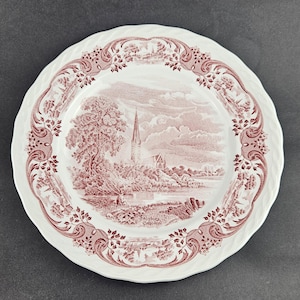 May include: A white ceramic plate with a scalloped edge featuring a maroon transferware print of Salisbury Cathedral. The central scene depicts the cathedral spire, trees, and a river, while the border displays ornate floral scrollwork patterns.