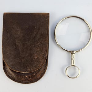 Peut inclure: Une loupe ronde avec un cadre en métal argenté et une poignée en forme d'anneau, placée à côté d'une pochette en cuir marron foncé texturé, dotée d'un rabat arrondi et de coutures visibles sur les bords.