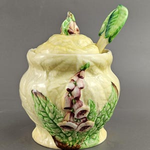 May include: A ceramic sugar bowl with a lid and spoon. The bowl is light yellow with a floral design of purple and green foxgloves. The spoon is green and white. The lid has a foxglove bud on top.