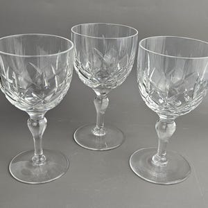 May include: Three clear crystal wine glasses with diamond-cut patterns. Each glass has a rounded bowl, a slender stem, and a circular base. The glasses are arranged on a grey surface, showcasing their elegant design.