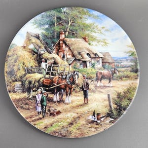 May include: A decorative plate featuring a pastoral scene with a thatched-roof cottage, hay wagon, horses, and figures. The artwork depicts a rural landscape with a hay harvest, conveying a sense of nostalgia and country life. The plate is round and has a glossy finish.