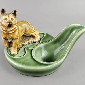 Könnte beinhalten: Ein Keramik-Aschenbecher mit einer Schäferhund-Figur in Braun- und Beigetönen. Der Aschenbecher hat eine blattförmige Form in einem gedämpften Grün mit einem gebogenen, länglichen Abschnitt. Ca. 20 cm lang.