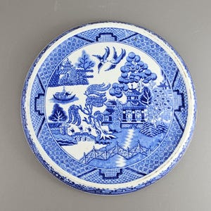 Könnte beinhalten: Runder blau-weißer Keramikteller mit Willow-Muster. Der Teller zeigt eine detaillierte Landschaftsszene mit Pagoden, Bäumen, einer Brücke und fliegenden Vögeln. Der Rand hat ein geometrisches Muster.