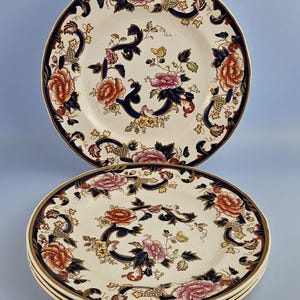 Könnte beinhalten: Ein Set aus vier runden Keramiktellern mit cremefarbener Basis und einem Blumenmuster in Rosa-, Orange- und Gelbtönen. Das Design umfasst dunkelblaue Schnörkelakzente und einen goldfarbenen Rand an der Kante jedes Tellers.