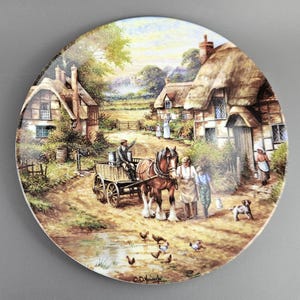 May include: Round decorative plate featuring a rural village scene with thatched-roof cottages, a horse-drawn cart, and figures in period clothing. The plate has a detailed, colorful illustration with a pastoral theme.