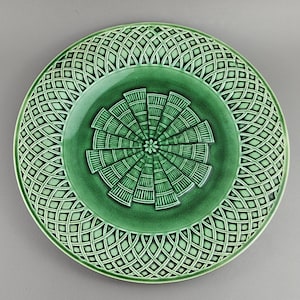 Puede incluir: Un plato de cerámica verde con un detallado patrón de celosía geométrica alrededor del borde. El centro presenta un diseño radial con un pequeño acento floral. El color del plato es un verde vibrante y sólido.