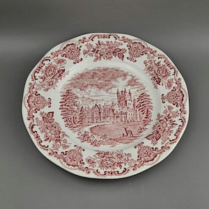 Puede incluir: Plato de cerámica blanca con borde floral rojo y escena central de un castillo y un ciervo. El plato tiene un borde festoneado y probablemente sea vintage o antiguo.