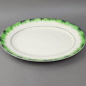 May include: An oval serving platter with a white centre and a green and black fringed border. A thin black line separates the white centre from the decorative edge. The platter is made of ceramic and is suitable for serving food.