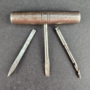Puede incluir: Una herramienta vintage con mango en T que presenta una empuñadura cilíndrica de metal desgastado y un eje central con punta de destornillador plano. Incluye dos accesorios metálicos desmontables: una hoja fina y puntiaguda y una barrena.