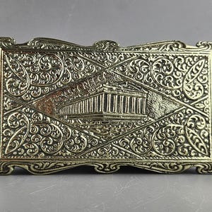 Puede incluir: Una caja decorativa rectangular dorada con intrincados patrones arremolinados y un diseño central en forma de diamante. El diseño presenta un relieve de un edificio con columnas, posiblemente un monumento histórico. La caja tiene un borde ornamentado.
