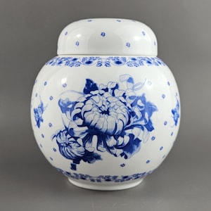 May include: A white ceramic jar with a lid, decorated with a large blue floral design. The jar has a round body and a matching lid with small blue dots. The floral pattern includes detailed blue flowers and leaves.