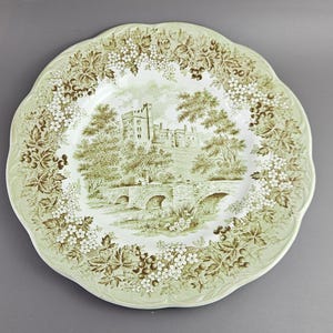 Puede incluir: Un plato redondo con borde festoneado, con un borde floral verde claro y blanco. El centro presenta un paisaje sepia de un castillo, un puente y árboles. El plato es probablemente de cerámica y es adecuado para servir comida.
