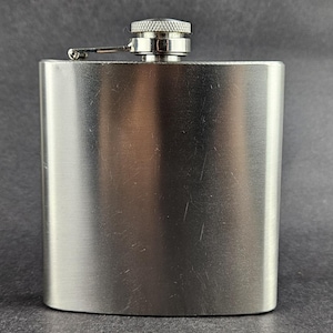 May include: A stainless steel hip flask with a brushed metal finish. The flask has a rounded rectangular shape with a screw-on cap and a hinged arm. The cap is silver and has a textured design. The flask is designed to hold liquids.