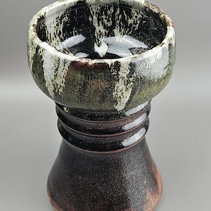 Può includere: Una ciotola in ceramica fatta a mano con base a piedistallo. Il bordo della ciotola presenta un motivo maculato nero, bianco e verde. Il piedistallo è marrone scuro con finitura strutturata. Adatta per servire o esporre.