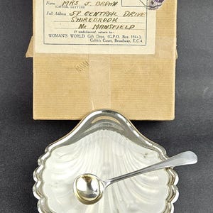 Puede incluir: Un plato plateado con forma de concha y una cuchara pequeña a juego, acompañado de una caja de cartón vintage para envíos. La caja tiene una etiqueta dirigida a Mrs. J. Brown en Shirebrook, Mansfield, con un sello postal de color morado.