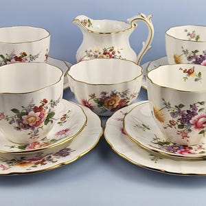 Może przedstawiać: Porcelanowy zestaw do herbaty składający się z czterech filiżanek, czterech spodków, czterech talerzyków, dzbanuszka na mleko i cukiernicy. Każdy element jest biały, z falistym brzegiem, złotym wykończeniem i kolorowym wzorem kwiatów i liści.