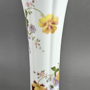 May include: White porcelain vase with a fluted design and a gold rim. The vase is decorated with colourful floral patterns, including yellow, purple, and pink flowers with green leaves, creating an elegant aesthetic.