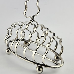 May include: A silver-colored, Art Deco-style toast rack. The rack has a handle and curved dividers, supported by small ball feet. The design is symmetrical and elegant, suitable for serving toast.