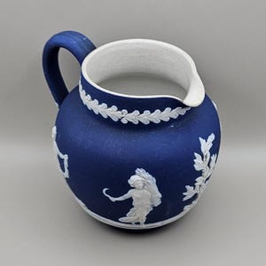 Puede incluir: Una jarra de cerámica azul con interior y asa blancos. La jarra presenta diseños en relieve blancos de figuras y follaje. Un borde decorativo blanco rodea la parte superior de la jarra.