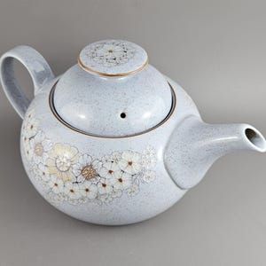 Könnte beinhalten: Eine hellblaue Keramik-Teekanne mit floralem Design. Die Teekanne hat einen abgerundeten Körper, einen gebogenen Griff und einen Ausguss. Der Deckel ist mit einem passenden Blumenmuster verziert. Die Teekanne ist mit weißen und gelben Blumen verziert.