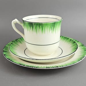 Puede incluir: Un juego de taza y platillo de té blanco con un diseño geométrico verde y negro. La taza tiene un asa y el platillo tiene un borde elevado.