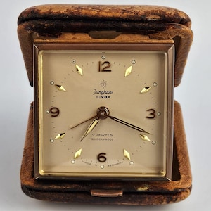 May include: A square Junghans Divox travel alarm clock with a cream-colored face, gold-toned hands, and gold-toned hour markers. The clock features the text Junghans, Divox, 7 Jewels, Shockproof, and Made in Germany, housed in a brown leather case.