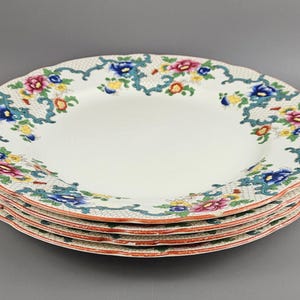 May include: A stack of white ceramic plates with a floral design. The plates have a scalloped edge and a red and white border. The floral pattern includes blue, pink, and yellow flowers with green vines.
