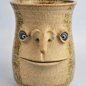 Puede incluir: Una taza cilíndrica de cerámica con acabado beige moteado que presenta un rostro esculpido. El rostro incluye una nariz prominente, una hendidura horizontal para la boca y dos ojos azules incrustados con pupilas oscuras a los lados.