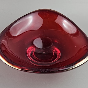 May include: A triangular, ruby-red glass bowl with a gold-coloured rim. The bowl has a smooth, glossy surface and a small, raised circle in the centre. The design is simple and elegant, suitable for decorative use.