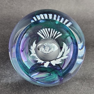 May include: A clear glass paperweight with a thistle design. The paperweight features a central sphere with an etched thistle, surrounded by a larger, clear glass orb with blue, green, and purple swirls. White accents highlight the thistle.