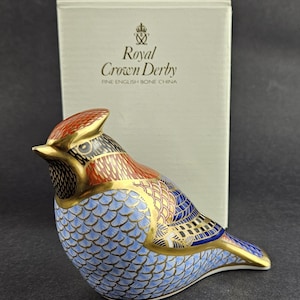 Puede incluir: Una figura de pájaro de porcelana fina inglesa Royal Crown Derby con una cresta en tonos naranja, negro y dorado. El cuerpo presenta un patrón de escamas en azul claro, dorado y azul oscuro. La caja tiene impreso el logo Royal Crown Derby.