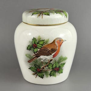 May include: A white ceramic jar with a lid, featuring a detailed illustration of a robin perched on a branch with holly. The lid has a gold rim and a matching bird and holly design. A decorative piece.