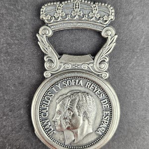 May include: Silver bottle opener with a crown design. The opener has a circular medallion with profiles of two people and the text "JUAN CARLOS I Y SOFIA REYES DE ESPAÑA."