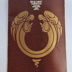 Peut inclure: Un programme souvenir et livret rectangulaire pour Jesus Christ Superstar avec une couverture marron foncé. Le devant présente un grand graphique doré stylisé de deux figures d'oiseaux se faisant face formant un cercle avec un symbole.