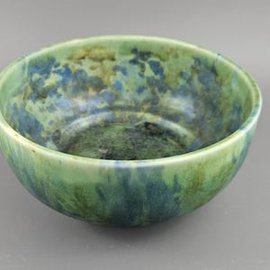 May include: A ceramic bowl with a speckled glaze in shades of green, blue, and brown. The bowl has a rounded shape and a smooth, glossy finish. The interior features darker hues, creating a contrast with the lighter exterior. A handcrafted item.